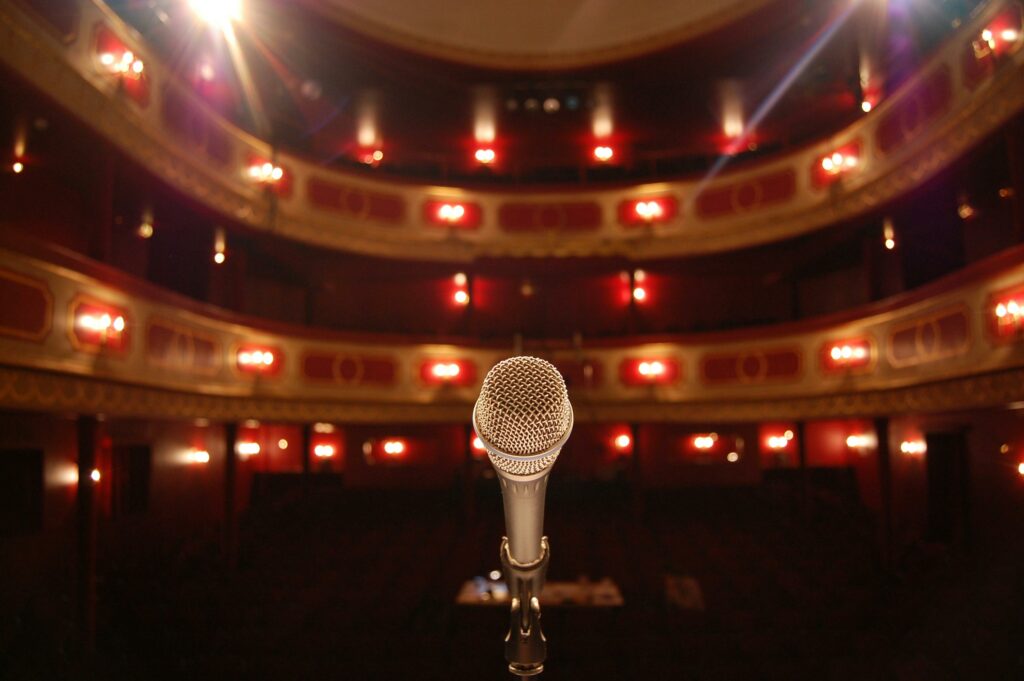 A microphone stands on a theater stage with ornate balconies and warm lighting.